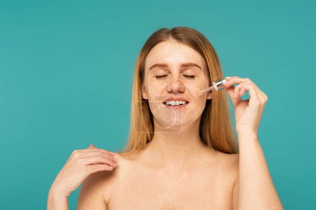 Smiling freckled woman applying serum on face isolated on turquoise