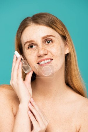 Carefree woman with freckles holding serum isolated on turquoise