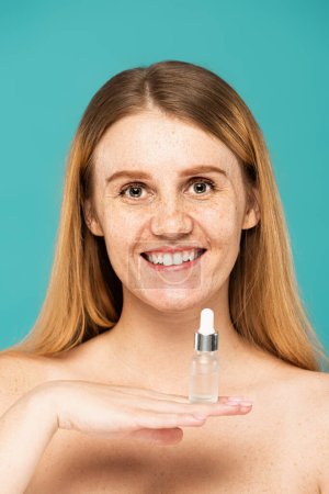 cheerful woman with freckles and red hair holding serum in bottle isolated on turquoise