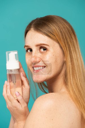 cheerful and redhead woman with freckles holding bottle with foam cleanser isolated on turquoise