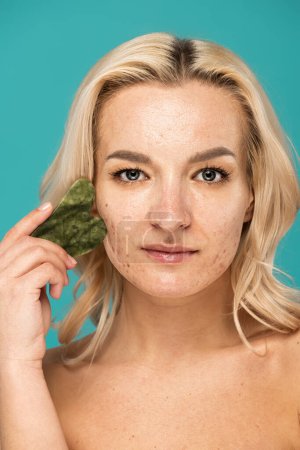 Téléchargez les photos : Femme blonde avec des imperfections massage visage avec grattoir visage isolé sur turquoise - en image libre de droit