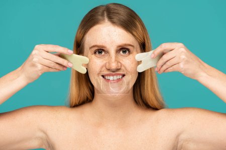 fröhliche Frau mit Sommersprossen mit Jade-Gesichtsschaber isoliert auf Türkis
