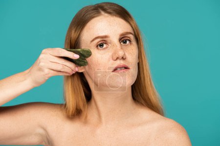 jeune et rousse femme sans maquillage en utilisant gua sha visage scrapper isolé sur turquoise
