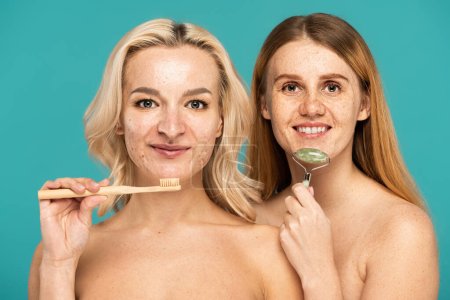 happy blonde woman brushing teeth near cheerful model using jade roller isolated on turquoise 