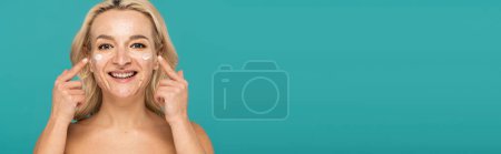 Téléchargez les photos : Femme gaie avec de l'acné appliquer la crème et pointant avec les doigts sur le visage isolé sur turquoise, bannière - en image libre de droit