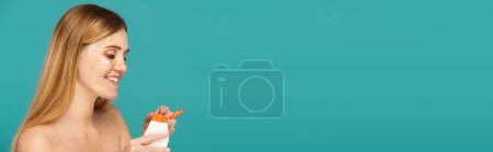 Téléchargez les photos : Femme rousse gaie avec des taches de rousseur tenant tube avec crème hydratante isolé sur turquoise, bannière - en image libre de droit