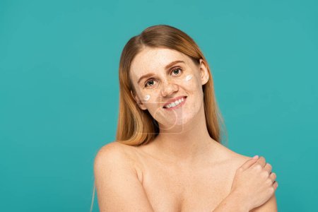 happy woman with freckles and cosmetic cream on cheeks isolated on turquoise