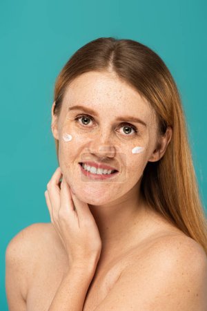smiling woman with freckles and cosmetic cream on face isolated on turquoise