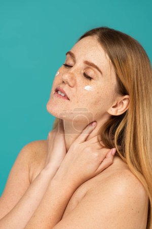 freckled woman with closed eyes and cosmetic cream on face isolated on turquoise