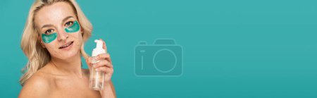 Téléchargez les photos : Femme blonde avec acné et patchs pour les yeux tenant bouteille avec nettoyant mousse isolé sur turquoise, bannière - en image libre de droit