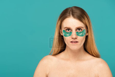 rousse femme avec des taches de rousseur et des taches de rousseur sous les yeux regardant la caméra isolée sur turquoise 