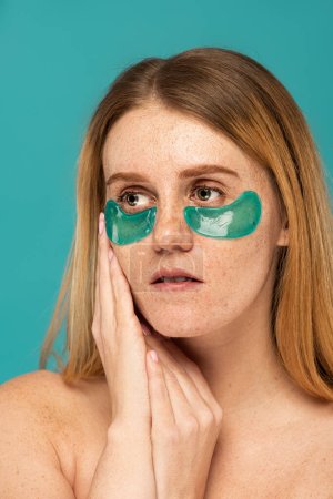 young woman with freckles and patches under eyes looking away isolated on turquoise 