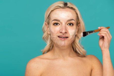 Photo for Blonde woman with acne holding cosmetic brush and applying clay mask isolated on turquoise - Royalty Free Image