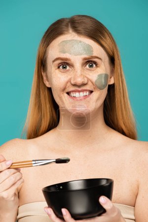 happy woman with red hair holding bowl and cosmetic brush while applying clay mask isolated on turquoise 