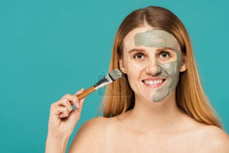 cheerful woman with red hair holding cosmetic brush while applying clay mask on half of face isolated on turquoise 