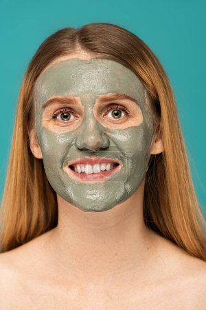 Téléchargez les photos : Gaie rousse femme avec masque d'argile sur le visage souriant isolé sur turquoise - en image libre de droit