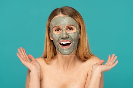 femme positive avec cheveux roux et masque d'argile sur le visage souriant isolé sur turquoise