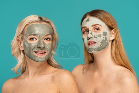 blonde and redhead women with clay mask on faces looking at camera isolated on turquoise