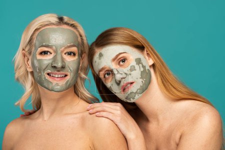 Foto de Rubia y pelirroja modelos con máscara de arcilla en las caras mirando a la cámara aislada en turquesa - Imagen libre de derechos