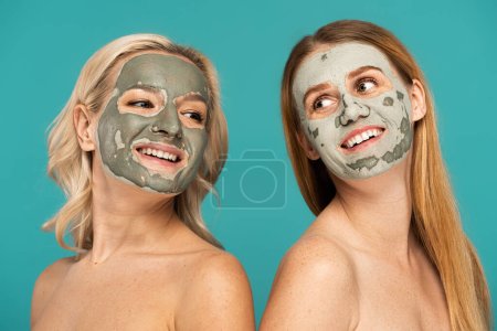 Photo for Cheerful redhead and blonde women with clay mask on faces posing isolated on turquoise - Royalty Free Image