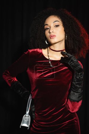 Fashionable african american woman in burgundy dress and gloves posing isolated on black 