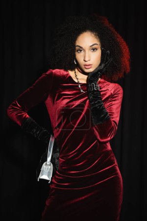 Trendiges afrikanisch-amerikanisches Model in Samtkleid und Handschuhen, das isoliert auf schwarz in die Kamera blickt 