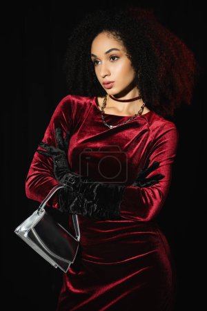 Femme afro-américaine élégante en robe posant avec sac à main isolé sur noir 