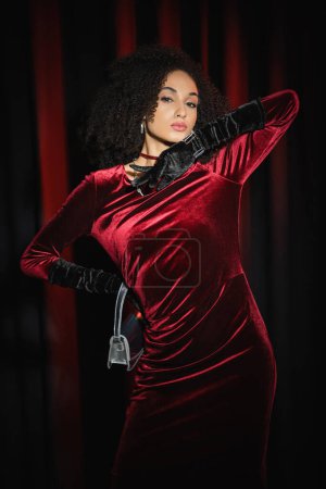 Foto de Modelo afroamericano de moda en vestido sosteniendo el bolso y de pie sobre fondo borgoña - Imagen libre de derechos