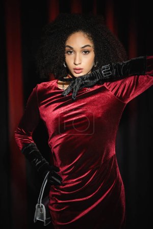 Hübsches afrikanisch-amerikanisches Model posiert in Samtkleid und Handschuhen neben weinrotem Vorhang 