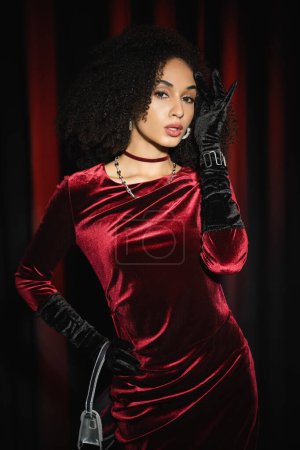 Pretty african american model posing in velvet dress and holding handbag near burgundy drapery 