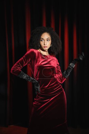 Stylish african american woman in gloves and dress posing near red drapery 