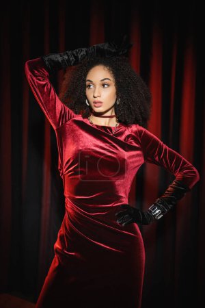 Elegante mujer afroamericana en guantes y vestido mirando hacia otro lado mientras posa cerca de cortinas rojas 