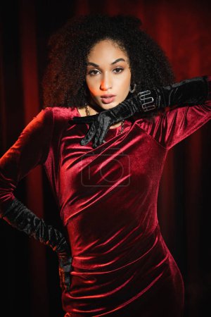 Fashionable african american woman in velvet dress and gloves posing near red drapery 