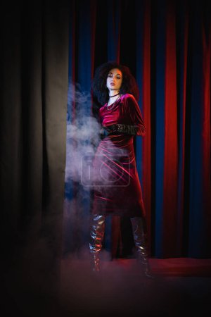 Foto de Joven modelo afroamericano en vestido borgoña de pie sobre fondo rojo con luz azul y humo - Imagen libre de derechos