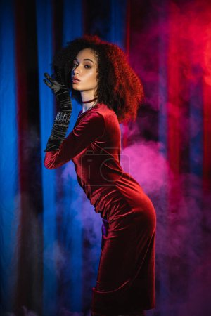 Stylish african american woman in velvet dress and glove posing near background with blue light and smoke
