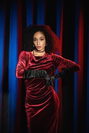 Modèle afro-américain bouclé en robe de velours et gants debout près de l'arrière-plan avec lumière bleue