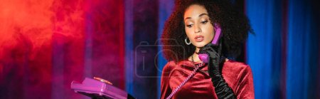 Foto de Elegante mujer afroamericana en guante de terciopelo hablando por teléfono sobre fondo rojo con luz azul con humo, pancarta - Imagen libre de derechos