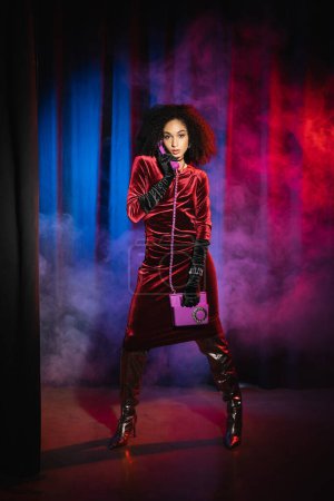 Foto de Elegante modelo afroamericano en vestido hablando por teléfono sobre fondo rojo con luz azul y humo - Imagen libre de derechos