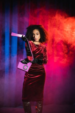 Foto de Modelo afroamericano de moda en vestido de terciopelo que sostiene el teléfono sobre fondo azul con luz roja y humo - Imagen libre de derechos