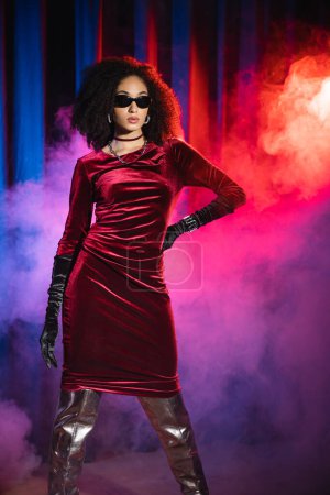 Stylish african american woman in sunglasses and velvet dress on red background with blue light and smoke