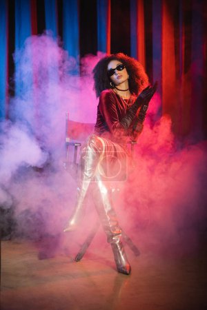 African american model in sunglasses and dress sitting on chair on red background with blue light and smoke 