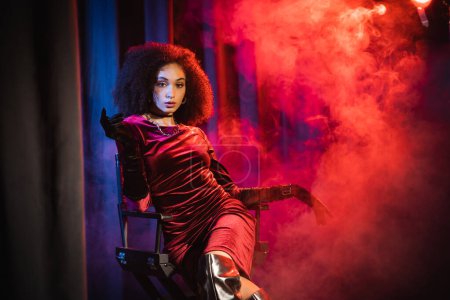 african american woman in velvet dress and gloves looking at camera while sitting on chair on background with red light and smoke