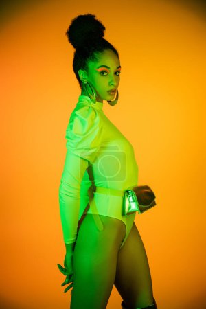 Stylish african american model with neon visage and bodysuit standing on orange background 