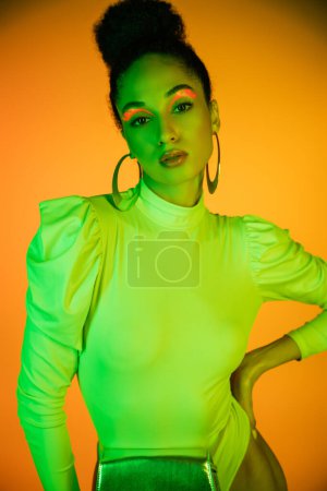 Stylish african american model with neon eyeliner and waist bag posing on orange background 