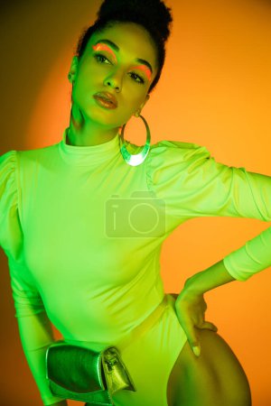 Fashionable african american model with neon visage posing in bodysuit on orange background  magic mug #648332606