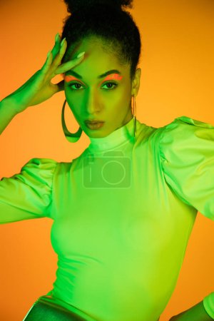 Portrait of fashionable african american model with neon visage touching face on orange background 