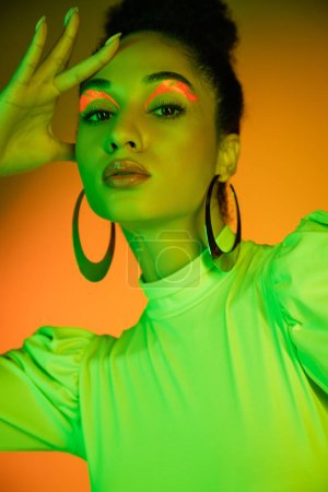Portrait of fashionable african american woman with neon visage standing on orange background 