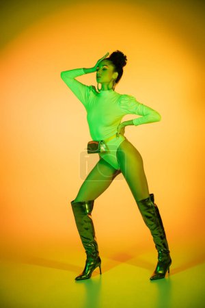 Trendy afrikanisch-amerikanische Frau in Neon-Body und Stiefeln posiert auf orangefarbenem Hintergrund 