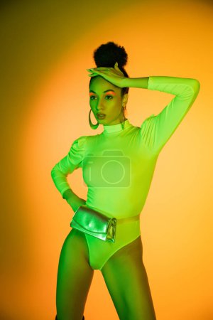 Stylish african american model in neon bodysuit touching hair on orange background 