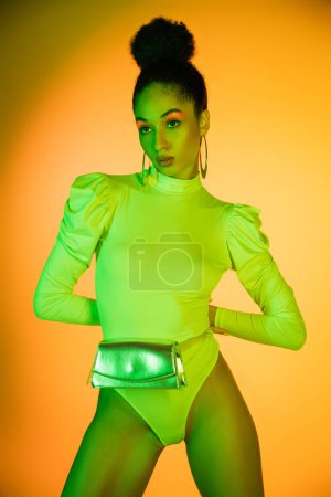 Pretty african american model in neon bodysuit and waist bag looking away on orange background 